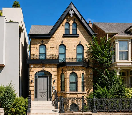 Restored yellow brick Victorian house with a steep gable, arched windows and black wrought-iron fence, home to Migize Pazaagwii Gamik in the Church-Wellesley Village